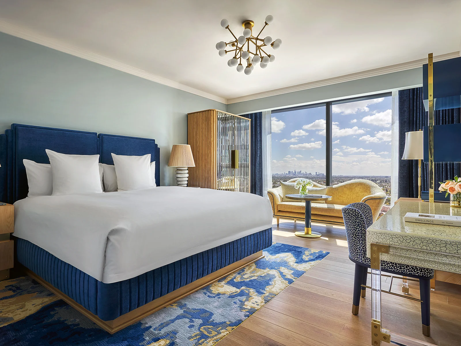 Hotel room with a blue bedframe and white sheets, and a picture window over blue sky and white clouds