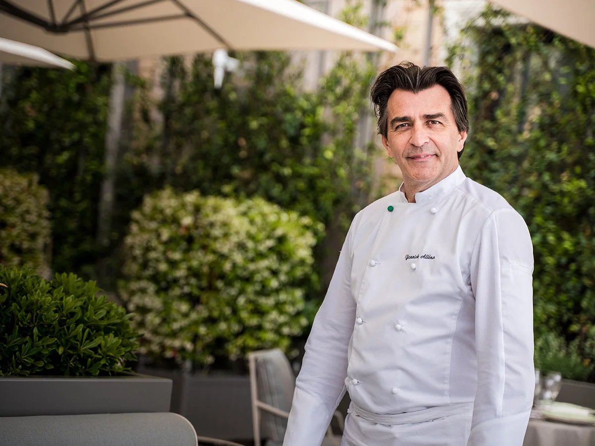 Chef Yannick Alléno ion his white uniform standing in the yard of a restaurant, with gray seats and hedges in tubs